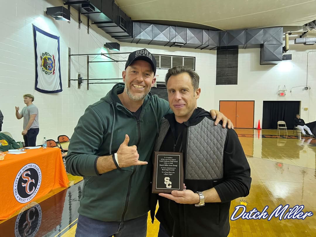 Two men smiling, one holding an award plaque, in a gymnasium setting.